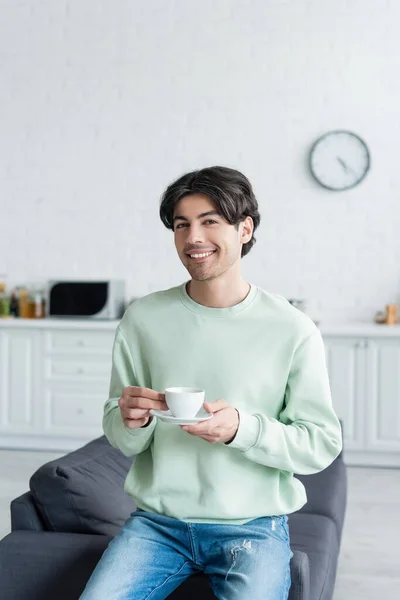 Homem alegre com xícara de café olhando para a câmera no sofá na cozinha embaçada — Fotografia de Stock