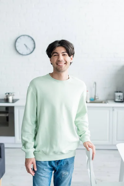 Young brunette man smiling at camera while standing in blurred kitchen — Stock Photo