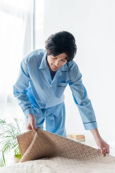 Sorrindo homem de pijama azul cobrindo cama com colcha de manhã — Fotografia de Stock