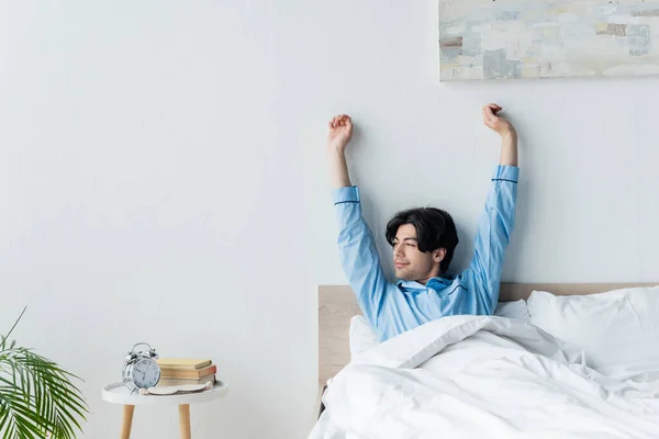 Улыбающийся мужчина растягивается с поднятыми руками, сидя в постели возле винтажного будильника — стоковое фото