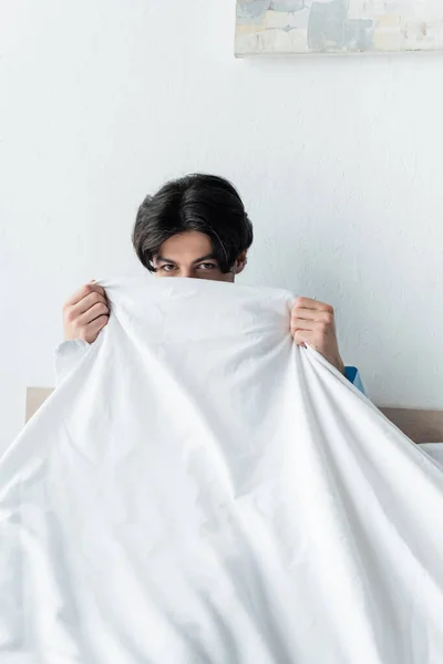 Brunette homme regardant caméra tout en obscurcissant visage avec couverture blanche — Photo de stock