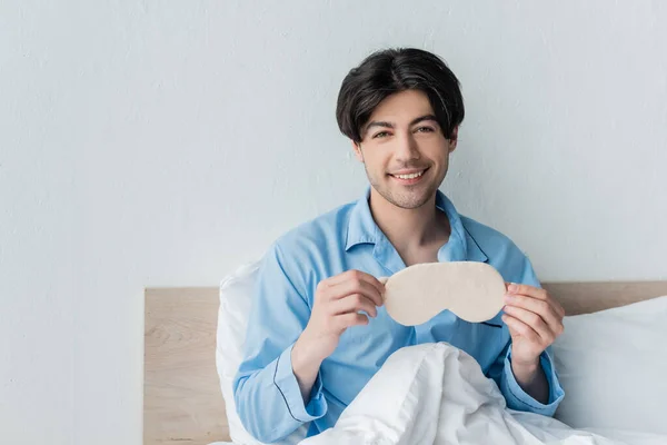 Happy man in blue pajamas sitting on bed with sleep mask — Stock Photo