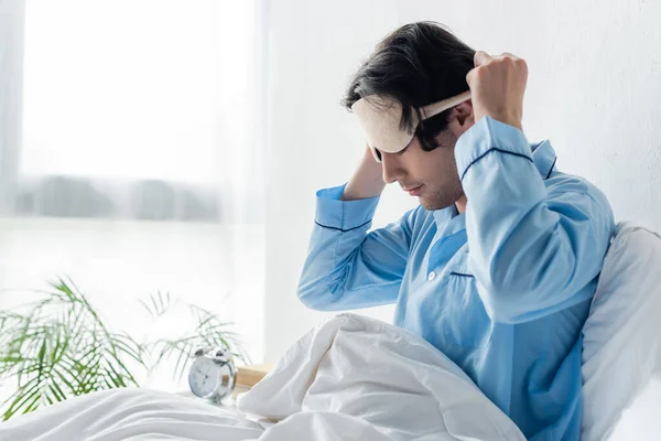 Hombre en pijama azul sentado en la cama por la mañana y quitándose la máscara del sueño - foto de stock