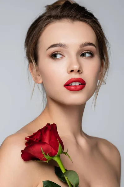 Mujer joven con labios rojos y piel perfecta mirando hacia otro lado cerca de rosa roja aislada en gris - foto de stock