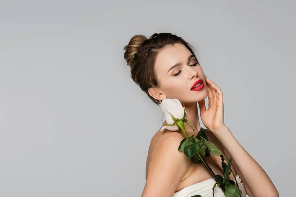 Bonita mujer con los ojos cerrados tocando la cara mientras sostiene rosa blanca aislada en gris - foto de stock