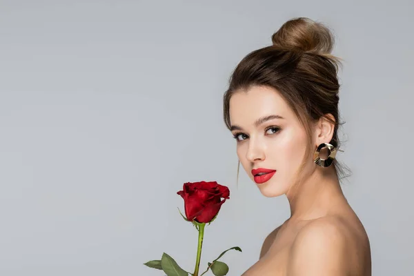 Jeune femme avec maquillage regardant caméra près de rose rouge isolé sur gris — Photo de stock