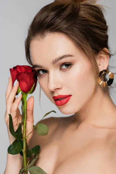 Vista de cerca de la encantadora mujer con maquillaje mirando a la cámara cerca de rosa roja aislado en gris - foto de stock