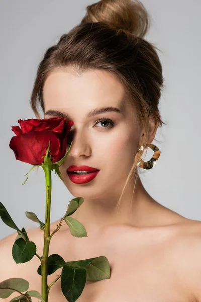 Affascinante donna con il trucco che tiene rosa rossa vicino al viso isolato sul grigio — Foto stock