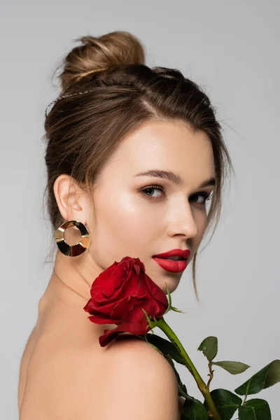 Hübsche junge Frau mit roten Lippen und Rose in der Nähe der nackten Schulter schaut in die Kamera isoliert auf grau — Stockfoto