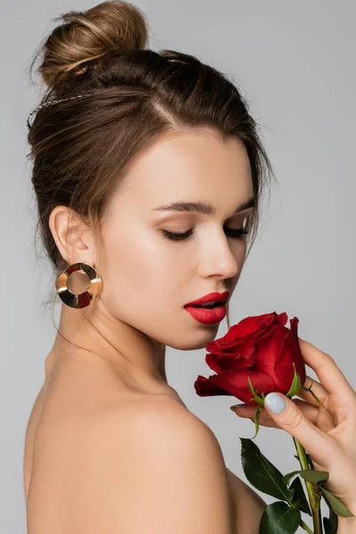 Bonita mujer en arete dorado sosteniendo rosa roja aislada en gris - foto de stock