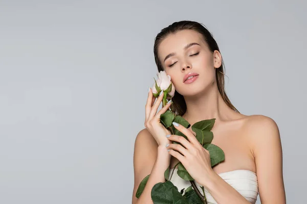 Mulher encantadora em top strapless posando com rosa branca e olhos fechados isolados em cinza — Fotografia de Stock