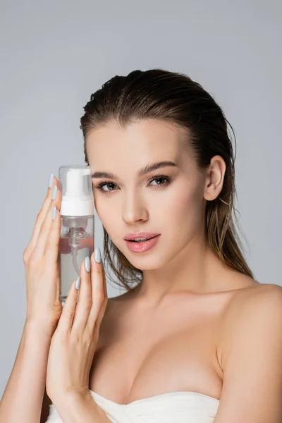 Junge Frau mit einer Flasche Gesichtsschaum blickt isoliert in die Kamera auf grau — Stockfoto