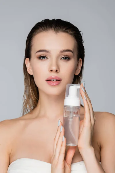 Jeune femme à la peau propre et aux cheveux mouillés tenant la mousse visage isolée sur gris — Photo de stock