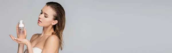 Jeune femme aux épaules nues et aux cheveux mouillés tenant la mousse visage isolée sur gris, bannière — Photo de stock
