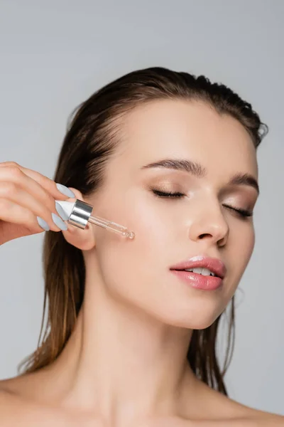 Close up view of charming woman with closed eyes applying cosmetic serum isolated on grey — Stock Photo