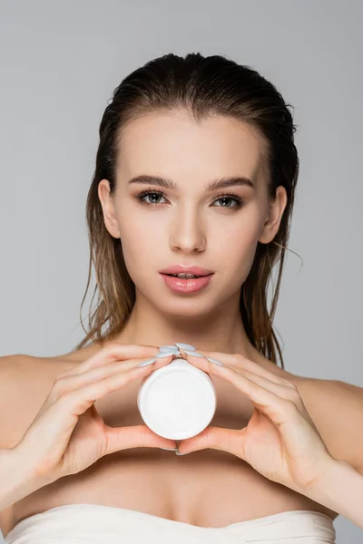 Pretty woman with container of cosmetic cream looking at camera isolated on grey — Stock Photo