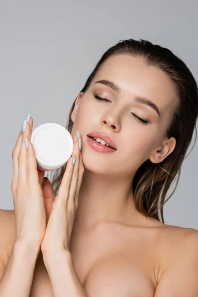 Mujer joven con el pelo mojado y los ojos cerrados celebración crema cosmética aislada en gris - foto de stock