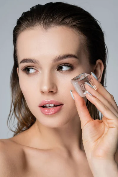 Vista da vicino della giovane donna con i capelli bagnati che tengono il cubetto di ghiaccio vicino al viso isolato sul grigio — Foto stock