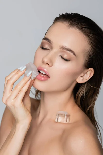 Mujer sensual con los ojos cerrados sosteniendo cubo de hielo cerca de los labios aislados en gris - foto de stock