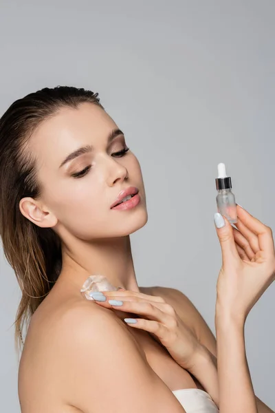 Pretty woman with ice cube on shoulder holding cosmetic serum isolated on grey — Stock Photo