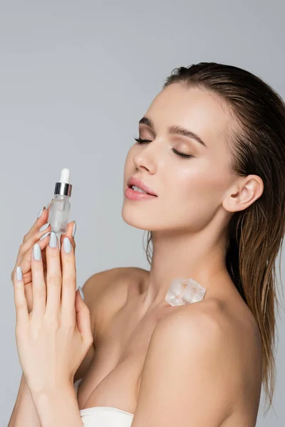 Sensual woman with wet hair and ice cube on body holding cosmetic serum isolated on grey — Stock Photo