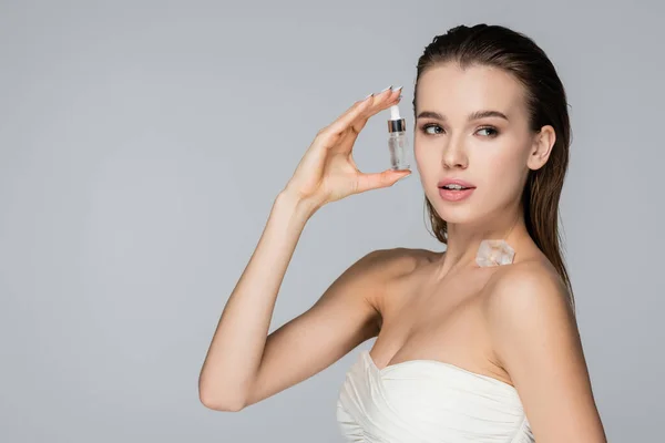 Young woman with cosmetic serum and ice cube on bare shoulder looking away isolated on grey — Stock Photo