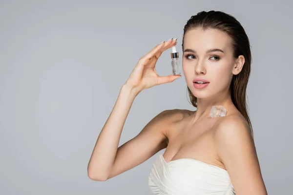 Mujer joven con suero cosmético y cubo de hielo en el hombro desnudo mirando hacia otro lado aislado en gris - foto de stock