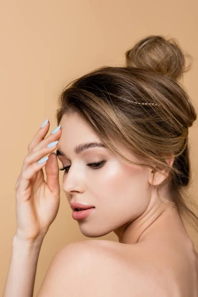Mujer joven con maquillaje natural y hombro desnudo tocando la frente aislada en beige - foto de stock