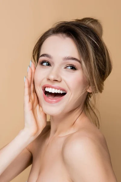 Mujer joven con maquillaje natural tocando la cara y riendo aislado en beige - foto de stock