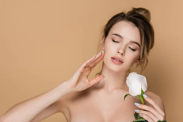 Jeune femme aux yeux fermés et aux épaules nues touchant le visage près de rose blanche isolée sur beige — Photo de stock