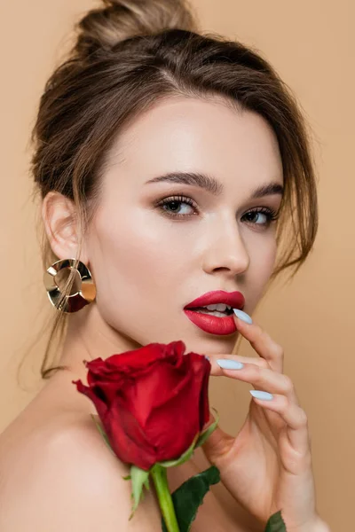 Vista de cerca de la joven mujer tocando los labios y mirando a la cámara cerca de rosa roja aislado en beige - foto de stock