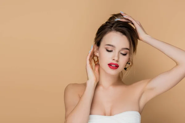 Pretty woman in strapless top and golden earrings fixing hair isolated on beige — Stock Photo