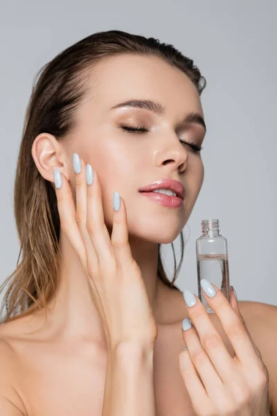 Mujer joven con el pelo mojado y ojos cerrados aplicando aceite cosmético aislado en gris - foto de stock