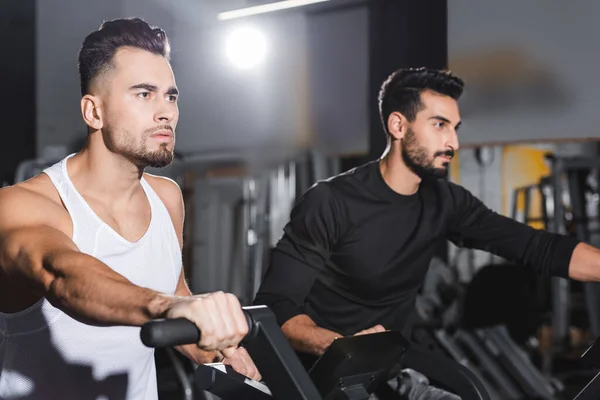 Young sportsman exercising on elliptical trainer near arabian man in gym — Stock Photo