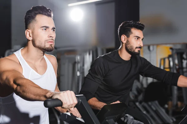 Joven deportista haciendo ejercicio en elíptica entrenador cerca de árabe hombre en el gimnasio - foto de stock