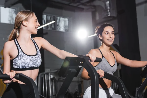 Cheerful interracial women training on elliptical trainers in sports center — Stock Photo