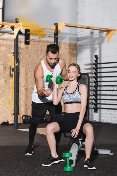 Mujer sonriente entrenando con pesas cerca del entrenador en el gimnasio - foto de stock