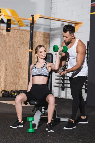 Mujer joven sonriente haciendo ejercicio con mancuerna cerca de entrenador en el centro deportivo - foto de stock