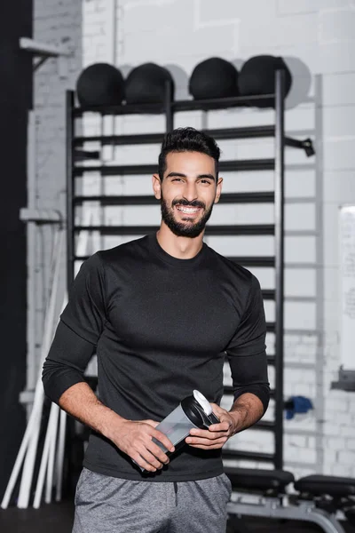 Alegre desportista árabe segurando garrafa de esportes e olhando para a câmera no ginásio — Fotografia de Stock