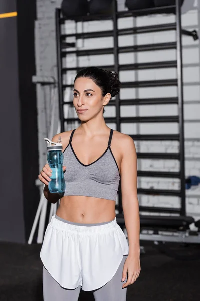 Sonriente deportista de Oriente Medio sosteniendo botella deportiva en el gimnasio - foto de stock