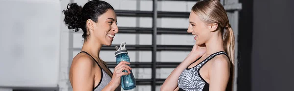 Vista lateral de la deportista sonriente hablando con un amigo árabe con botella de deporte en el gimnasio, pancarta - foto de stock