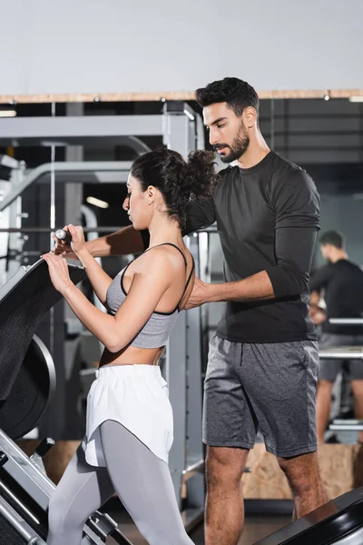 Arabian trainer standing near middle east sportswoman and leg press machine in gym — Stock Photo