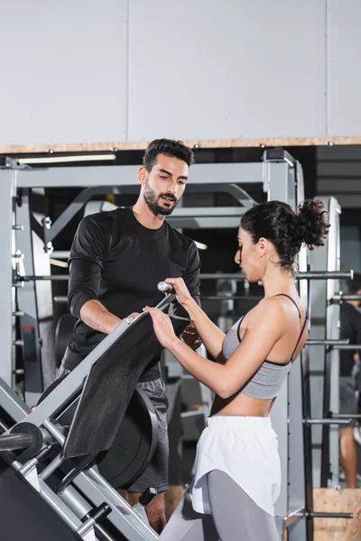 Nahost-Sportlerin steht neben Beinpresse und muslimischem Trainer im Fitnessstudio — Stockfoto