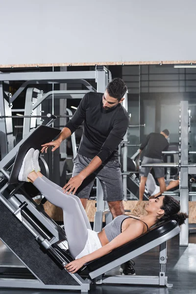 Middle east sportswoman training on leg press machine near arabian trainer in gym — Stock Photo