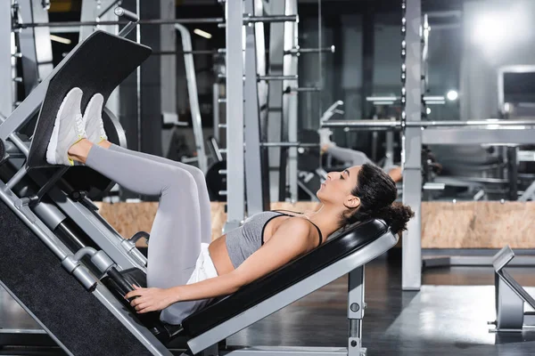 Vista laterale della sportiva mediorientale che si esercita sulla pressa per le gambe in palestra — Foto stock