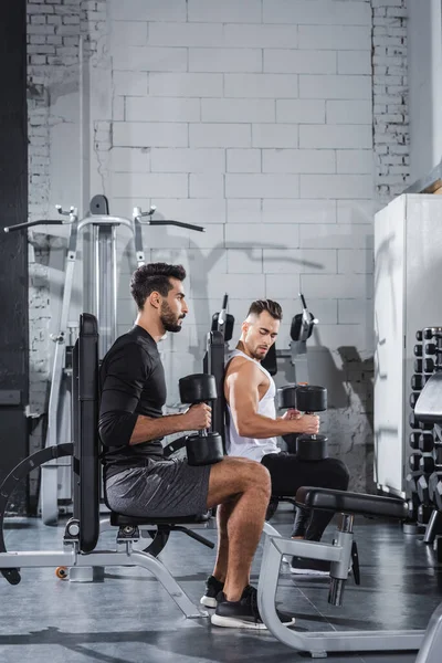 Young interracial sportsmen training with dumbbells and sports equipment in gym — Stock Photo