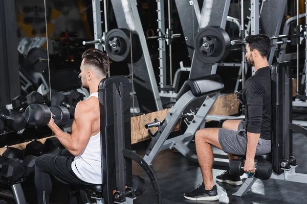 Multiethnic athletic sportsmen training with dumbbells near sports equipment in gym — Stock Photo