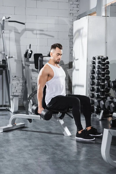 Athletic sportsman working out with dumbbell near sports equipment in gym — Stock Photo