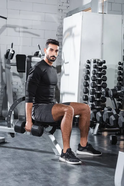 Joven deportista árabe mirando a la cámara mientras sostiene la mancuerna en el gimnasio - foto de stock