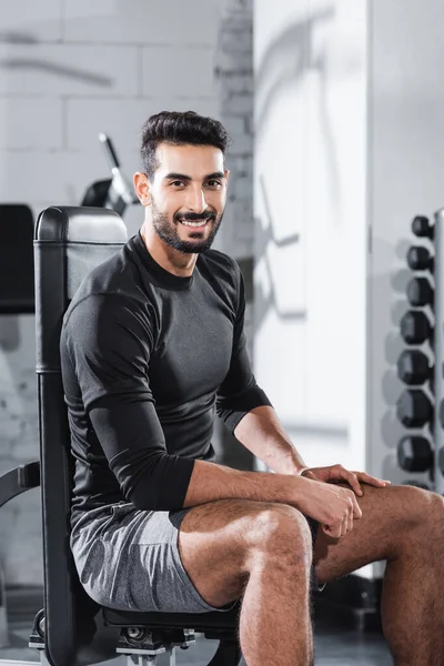 Sportif arabe souriant regardant la caméra tout en étant assis sur un équipement sportif dans la salle de gym — Photo de stock